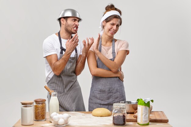 Ama de casa molesta no quiere escuchar al marido molesto, cocinar la cena juntos, permanecer irritada y cansada, usar productos saludables, hacer masa, aislado en la pared blanca. Culinaria, comida y gente