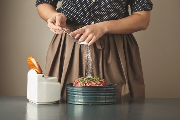 Ama de casa mujer irreconocible agrega un poco de sal en la carne picada en un recipiente de cerámica sobre la mesa azul