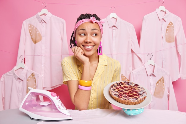 Ama de casa multitarea de piel oscura complacida hornea pastel y plancha la ropa para la familia usa pulseras de diadema posa cerca de la tabla de planchar sonríe feliz posa contra la pared rosa