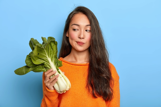 Ama de casa pensativa sostiene bok choy verde, piensa qué cocinar con esta verdura útil, sigue la dieta, es vegetariana, usa un jersey naranja