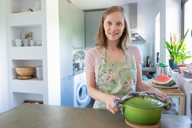 Ama de casa positiva cocinando en su cocina, sosteniendo una cacerola caliente con una toalla, mirando a la cámara y sonriendo. Concepto de cocina en casa