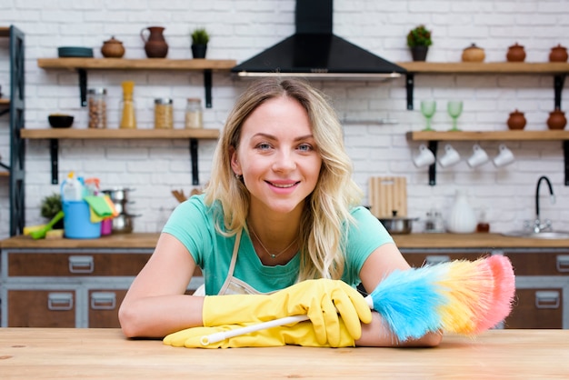 Ama de casa sonriente apoyándose en el mostrador de la cocina con plumero mirando a la cámara