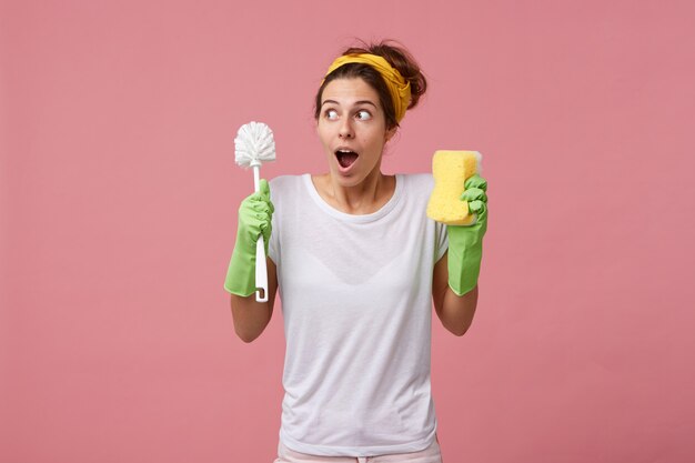 Ama de casa sorprendida en camiseta blanca informal y guantes protectores para limpiar yendo a ordenar la habitación sosteniendo un cepillo con una esponja con mirada perpleja mientras recuerda su reunión con un amigo