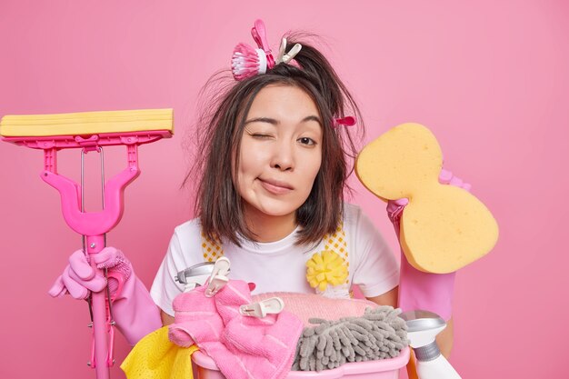 El ama de casa tiene el cabello desordenado, guiña los ojos, los ojos le proporcionan servicio de limpieza, sostiene una esponja y un trapeador limpios, siendo un limpiador profesional ocupado con las tareas del hogar