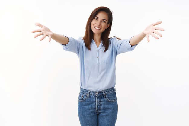 Ama de casa tierna y amigable extendió las manos a los lados agradablemente, sonriendo alegremente, invitando a amigos a entrar, sonriendo abiertamente esperando un abrazo, abrazos románticos, de pie en la pared blanca optimista, bienvenida a los invitados