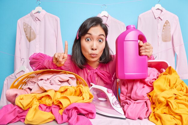 Ama de llaves asiática mantiene el dedo índice levantado sostiene la botella de detergente para planchar la ropa después del lavado obtiene una excelente idea dedica mucho tiempo al trabajo doméstico