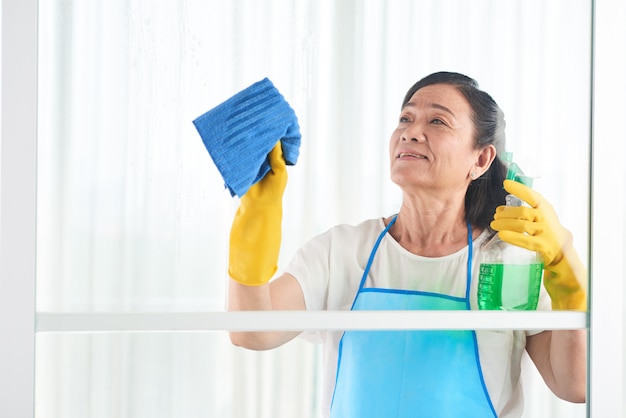 Ama de llaves de mediana edad limpiando la ventana con limpiador en aerosol