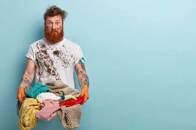 Ama de llaves sucia desconcertada viste una camiseta blanca informal, sostiene una canasta con ropa de cama, lava la ropa durante el fin de semana