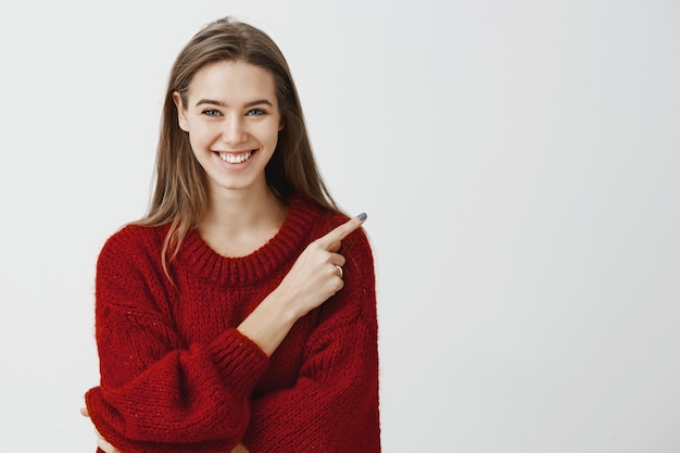 Amable y amable asistente de tienda listo para ayudar a encontrar el camino. Retrato de atractiva mujer europea alegre en suéter rojo suelto, apuntando a la esquina superior derecha, sonriendo ampliamente y expresando un estado de ánimo positivo