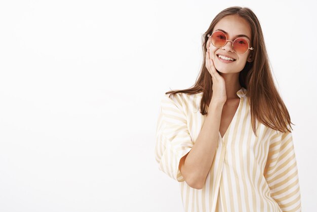 Amable atractivo y elegante joven sociable con cabello castaño con gafas de sol rojas y blusa blanca de verano tocando la cara y sonriendo