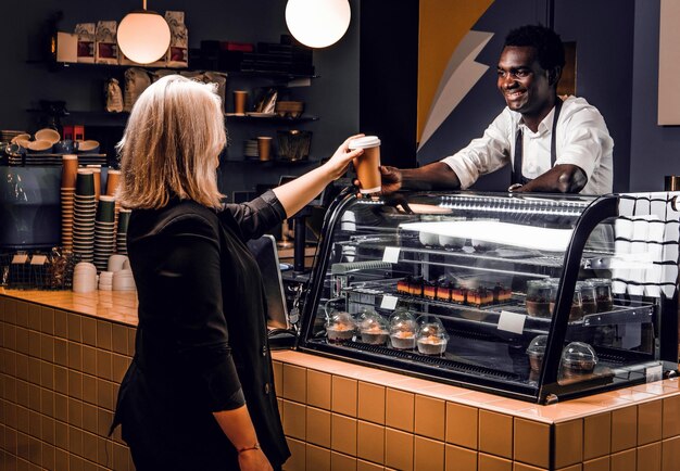 El amable barista africano le da al cliente café pedido de una cafetería moderna