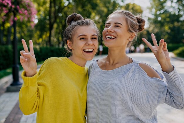 Amable chica sonriente en suéter amarillo disfrutando de una sesión de fotos al aire libre con su mejor amiga Impresionante modelo de cabello oscuro abrazando a su hermana en la calle