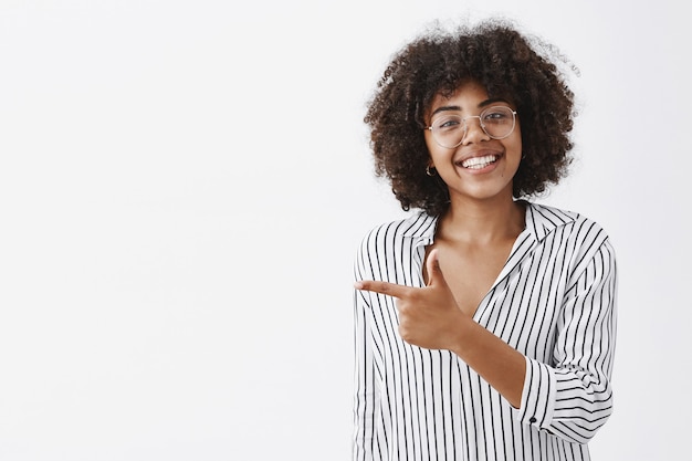 Amable y educada chica afroamericana de buen aspecto con gafas con el pelo rizado en una blusa de oficina a rayas apuntando hacia la izquierda con la pistola de dedo y sonriendo con alegría dando consejos a dónde ir