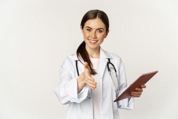 Amable joven doctora, médico estira la mano para el apretón de manos, saluda al paciente en la clínica, sostiene una tableta digital con datos médicos, fondo blanco.