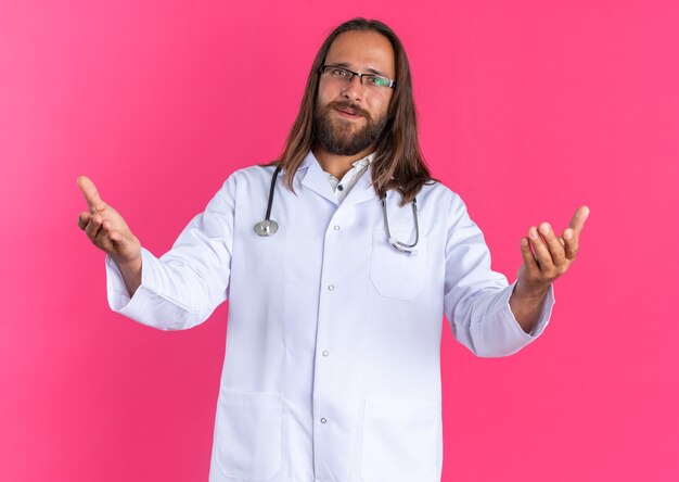 Amable médico varón adulto vistiendo bata médica y estetoscopio con gafas haciendo gesto de bienvenida