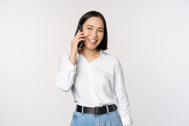 Amable mujer asiática sonriente hablando por teléfono chica de guardia sosteniendo el teléfono inteligente y riendo hablando de pie sobre fondo blanco.
