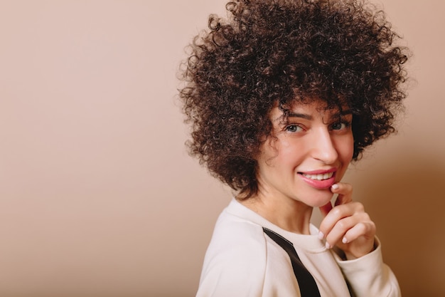 Amable mujer linda con rizos y maquillaje desnudo sonríe a la cámara y disfruta de buen humor en beige