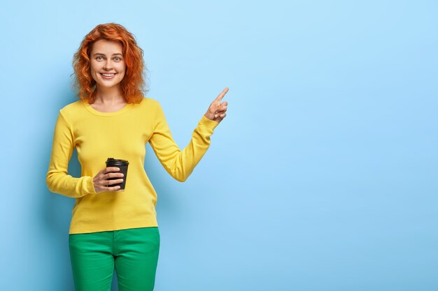 Amable mujer pelirroja sostiene una taza desechable de capuchino, muestra el camino a la cafetería, viste ropa de moda