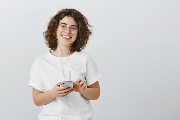 Amable mujer sonriente en vasos sosteniendo teléfono móvil, escuchando música en auriculares