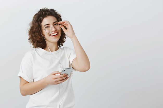 Amable mujer sonriente en vasos sosteniendo teléfono móvil, escuchando música en auriculares