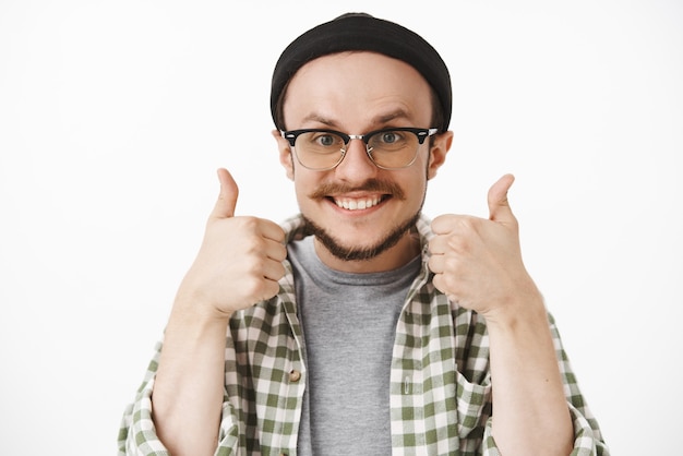 Foto gratuita amable optimista y feliz guapo hombre con barba y bigote en gorro negro y gafas transparentes sonriendo con alegría mostrando los pulgares hacia arriba en señal de aprobación o como gesto