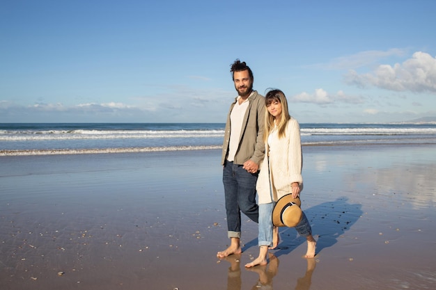 Amable pareja caminando cerca del agua en verano. hombre y mujer barbudos con ropa informal cogidos de la mano, paseando por la playa, mirando la cámara. amor, viajes, concepto de citas.