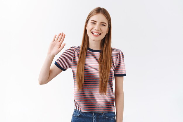 Amable positivo, linda joven caucásica en camiseta a rayas levantando la palma, saludando con la mano saluda, hola, saludando a un compañero de clase, sonriendo alegre y relajado, dando la bienvenida a los invitados, de pie fondo blanco