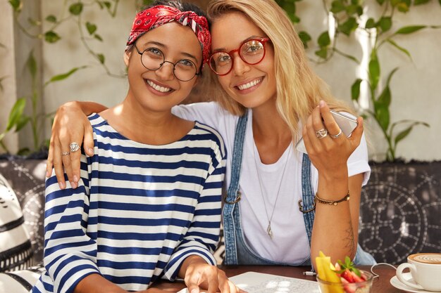 Amables mujeres de raza mixta con expresión positiva, se abrazan mientras pasan el tiempo libre en la cafetería, usan gafas