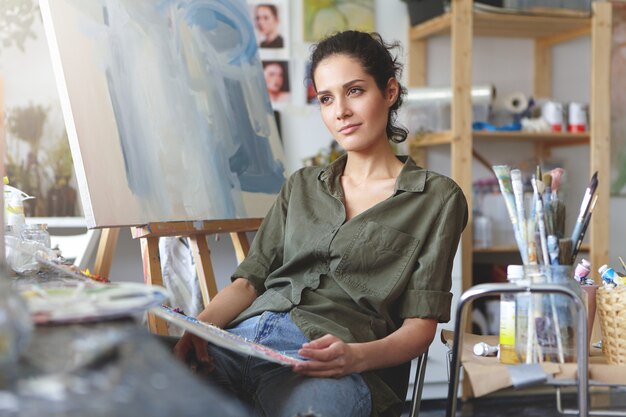 Amante del arte femenino haciendo bocetos preliminares, tratando de imaginar su futura obra maestra, teniendo una expresión pensativa rodeada de reproducciones de arte.