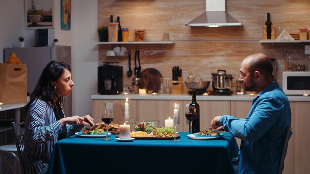 Amantes cenando juntos, comiendo y bebiendo vino durante la cena festiva en la cocina. Feliz pareja hablando, sentado a la mesa disfrutando de la comida en casa pasando un momento romántico juntos luces de velas sorpresa