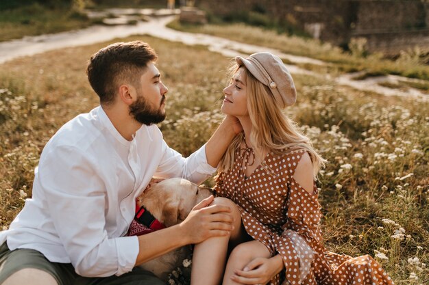 Los amantes se miran suavemente a los ojos, sentados en el parque sobre el césped junto al perro. El hombre acaricia suavemente a su novia.