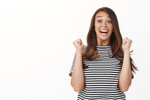 Foto gratuita ambiciosa, afortunada y esperanzada, linda chica ganadora de la lotería, saltando de la felicidad y la alegría, sintiéndose aliviada y optimista, con el puño de la cámara sonriendo y mirando asombrada, triunfante pared blanca