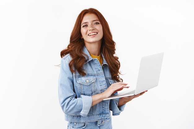 Ambiciosa, entusiasta, guapa, chica líder en vanlife, trabajando a distancia, de pie sobre fondo blanco, elegante chaqueta de mezclilla, sostenga una computadora portátil, sonriendo, satisfecha, trabajando independientemente, escribiendo un ensayo universitario.