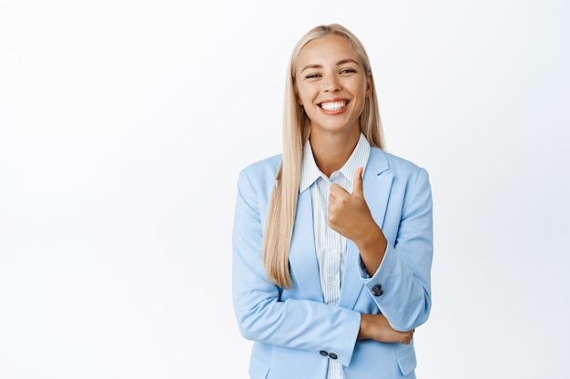 Ambiciosa mujer corporativa sonriente en traje que muestra los pulgares hacia arriba recomendando algo bueno de pie sobre fondo blanco.