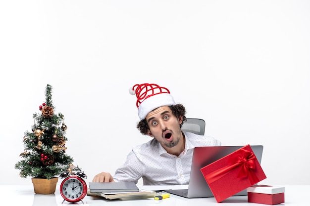 Ambiente festivo de vacaciones con empresario sorprendido con sombrero de santa claus trabajando solo en el proyecto en la oficina sobre fondo blanco.