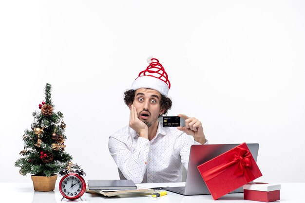 Ambiente festivo de vacaciones con un joven empresario barbudo sorprendido con sombrero de santa claus sosteniendo una tarjeta bancaria y poniendo su mano debajo de la barbilla en la oficina