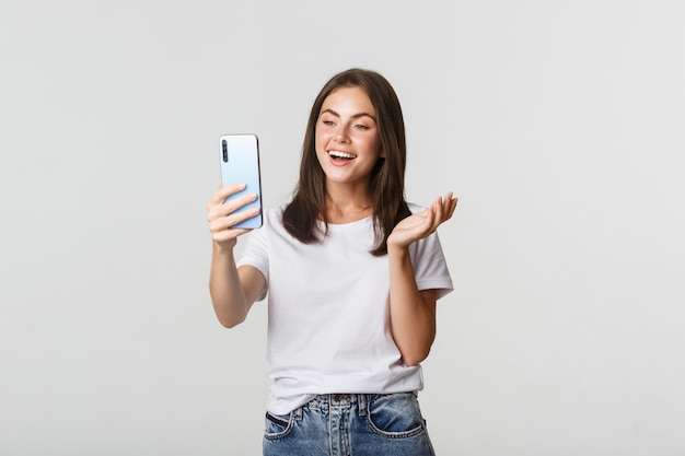 Amiga de videollamadas amigable chica atractiva, sonriendo y conversando, sosteniendo el teléfono inteligente, blanco.