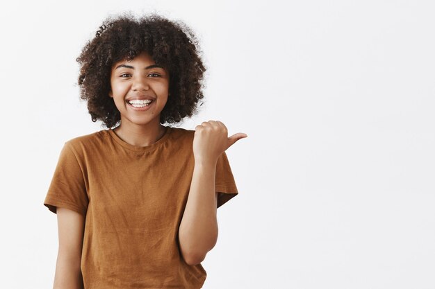 Amigable, atractiva, optimista, adolescente de piel oscura con peinado afro y elegante camiseta marrón apuntando a la derecha con el pulgar y sonriendo ampliamente mientras da buenos consejos