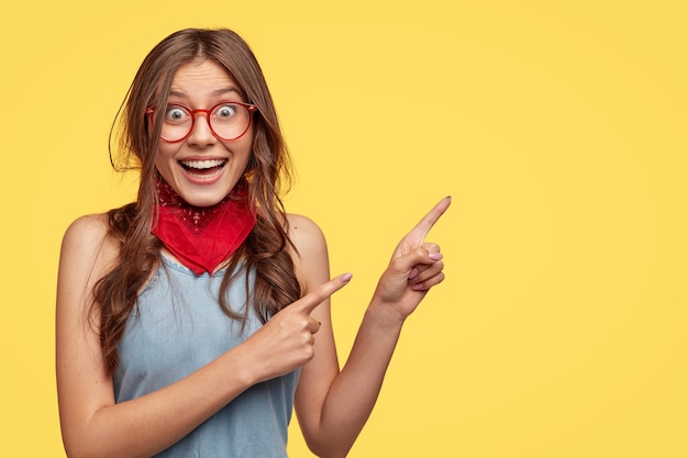 Foto gratuita la amigable y despreocupada asistente de tienda apunta a la derecha, tiene una sonrisa en el tablero, anuncia un nuevo atuendo con grandes descuentos, usa lentes transparentes, modelos contra la pared amarilla con espacio para copiar el lema