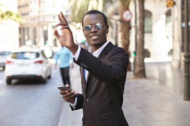 Amigable y exitoso joven empresario afroamericano con elegante traje negro y gafas enviando mensajes de texto por teléfono celular y levantando la mano mientras tomaba un taxi, parado en la calle en un entorno urbano