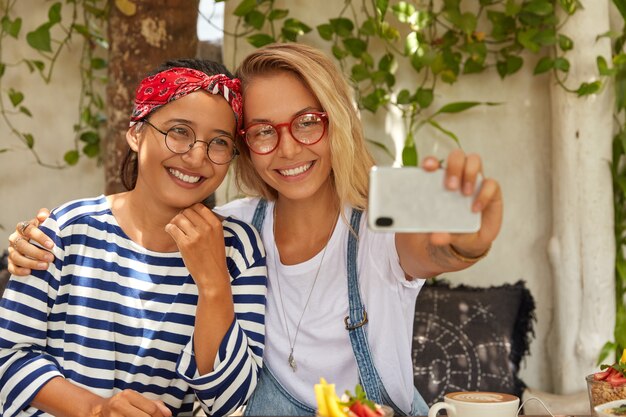 Amigables mujeres multiétnicas se abrazan y posan en el teléfono celular, hacen retratos autofotos, pasan el pasatiempo en la cafetería, comen postre, usan gafas redondas, disfrutan del tiempo de recreación, están satisfechas con algo