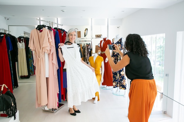 Amigas emocionadas disfrutando juntos de compras en la tienda de moda, sosteniendo el vestido y tomando fotos en el teléfono móvil. Consumismo o concepto de compras