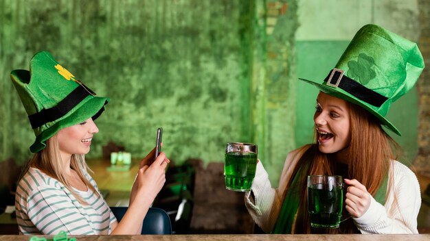 Amigas felices celebrando st. día de patricio junto con teléfono inteligente y bebidas.