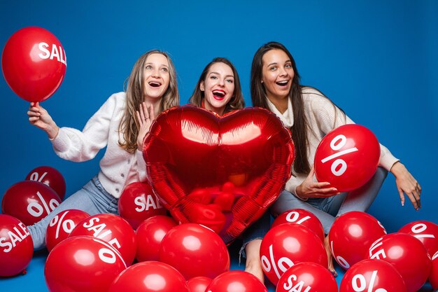 Amigas felices posando con globos rojos en forma de corazón y bolas de aire con letras de porcentaje y venta sobre fondo azul