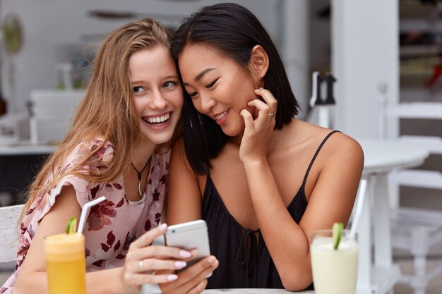 Las amigas jóvenes de raza mixta leen la notificación en el teléfono inteligente, tienen expresiones faciales positivas, beben jugo fresco,