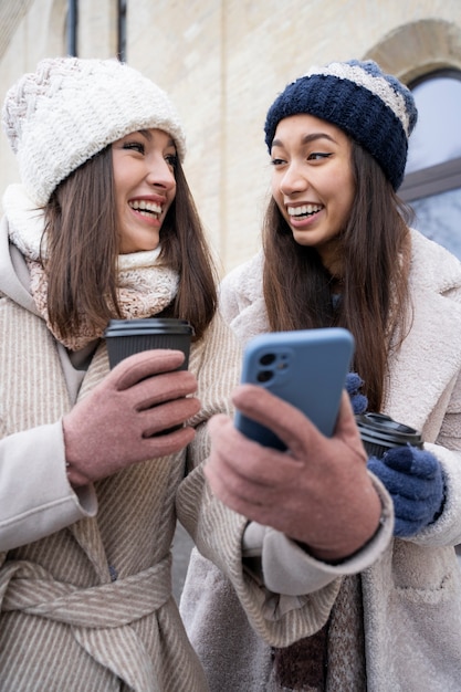 Foto gratuita amigas mirando el teléfono inteligente y sosteniendo tazas de café al aire libre después de reunirse