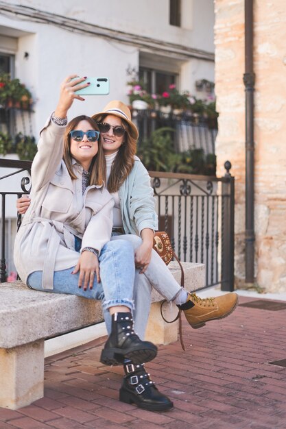 Amigas de moda tomando un selfie mientras está sentado en un banco en la calle