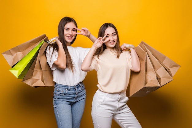 Las amigas de las mujeres jóvenes emocionadas sostienen la bolsa del paquete con las compras después de ir de compras posando aisladas en la pared amarilla. Concepto de estilo de vida de personas.