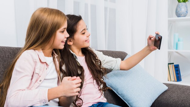 Amigas sentadas en el sofá tomando selfie en teléfonos inteligentes