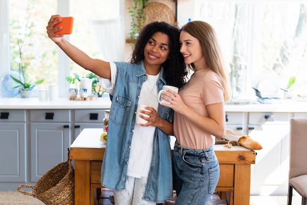 Amigas sonrientes tomando selfie en la cocina con smartphone
